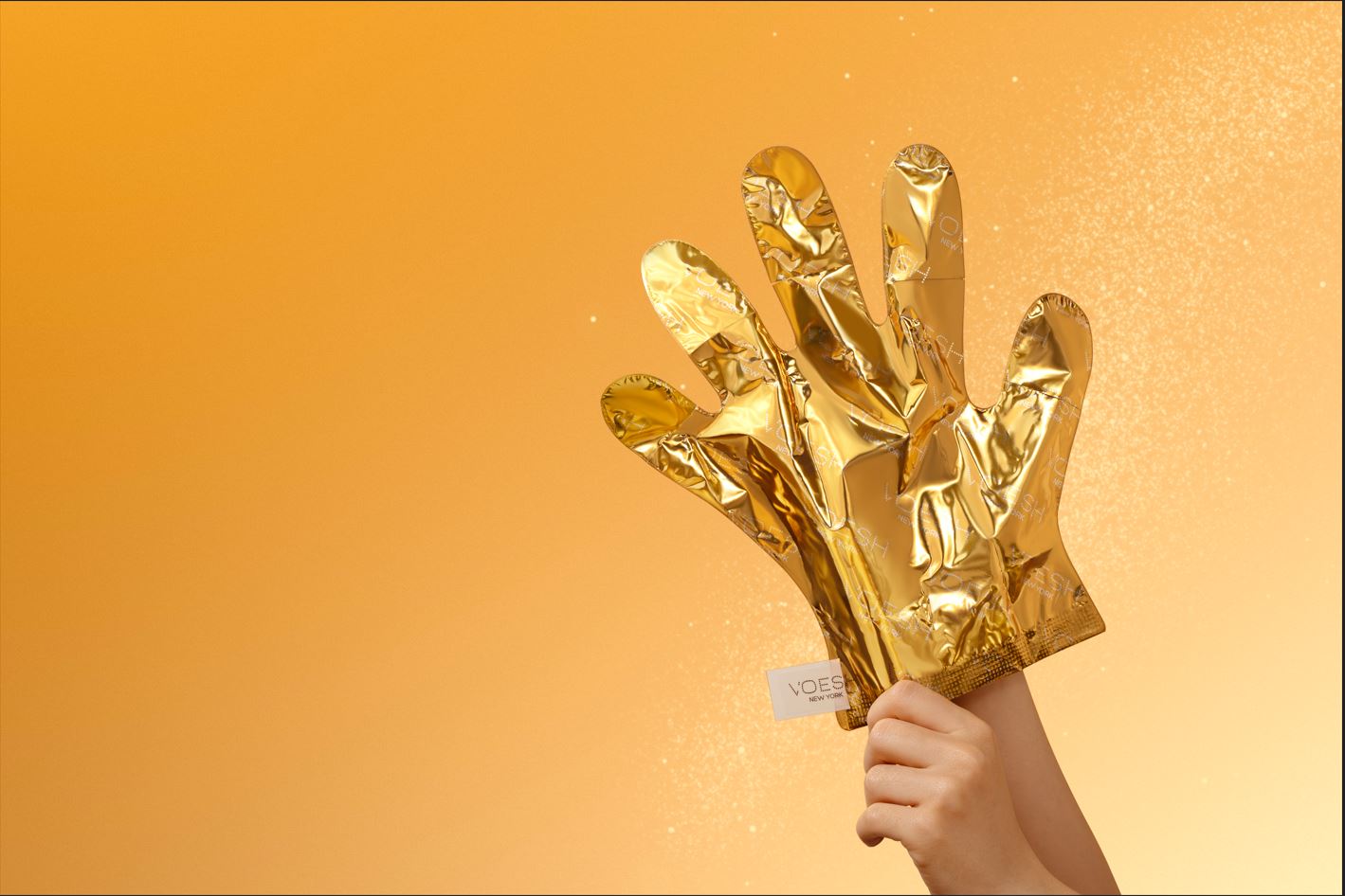 Eine Hand hält zwei goldene Handmasken von VOESH New York vor einem goldfarbenen, leuchtenden Hintergrund.