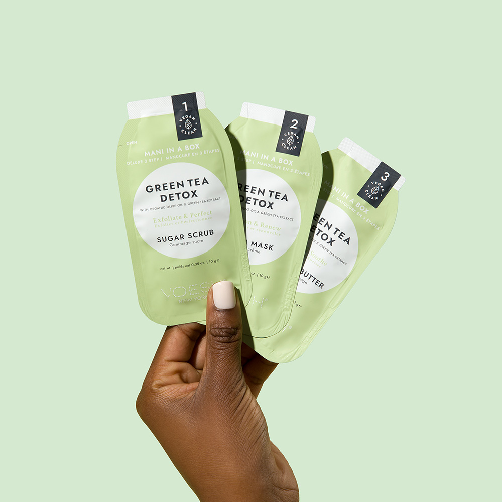 Hand hält drei Sachets des Voesh Mani in a Box Green Tea Detox Maniküre-Sets mit Sugar Scrub, Mask und Butter vor hellgrünem Hintergrund
