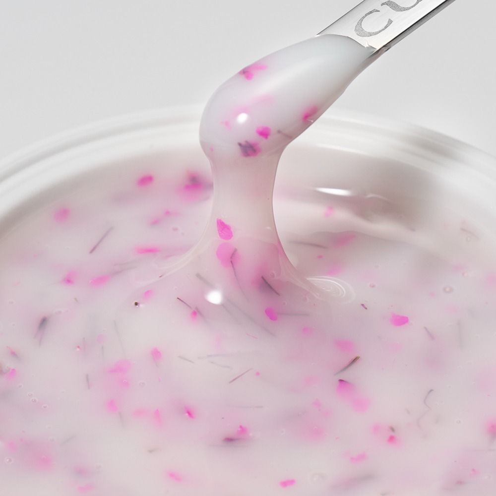 Nahaufnahme eines cremig-rosa Pflegegels mit pinken Blütenpartikeln auf einem Spatel