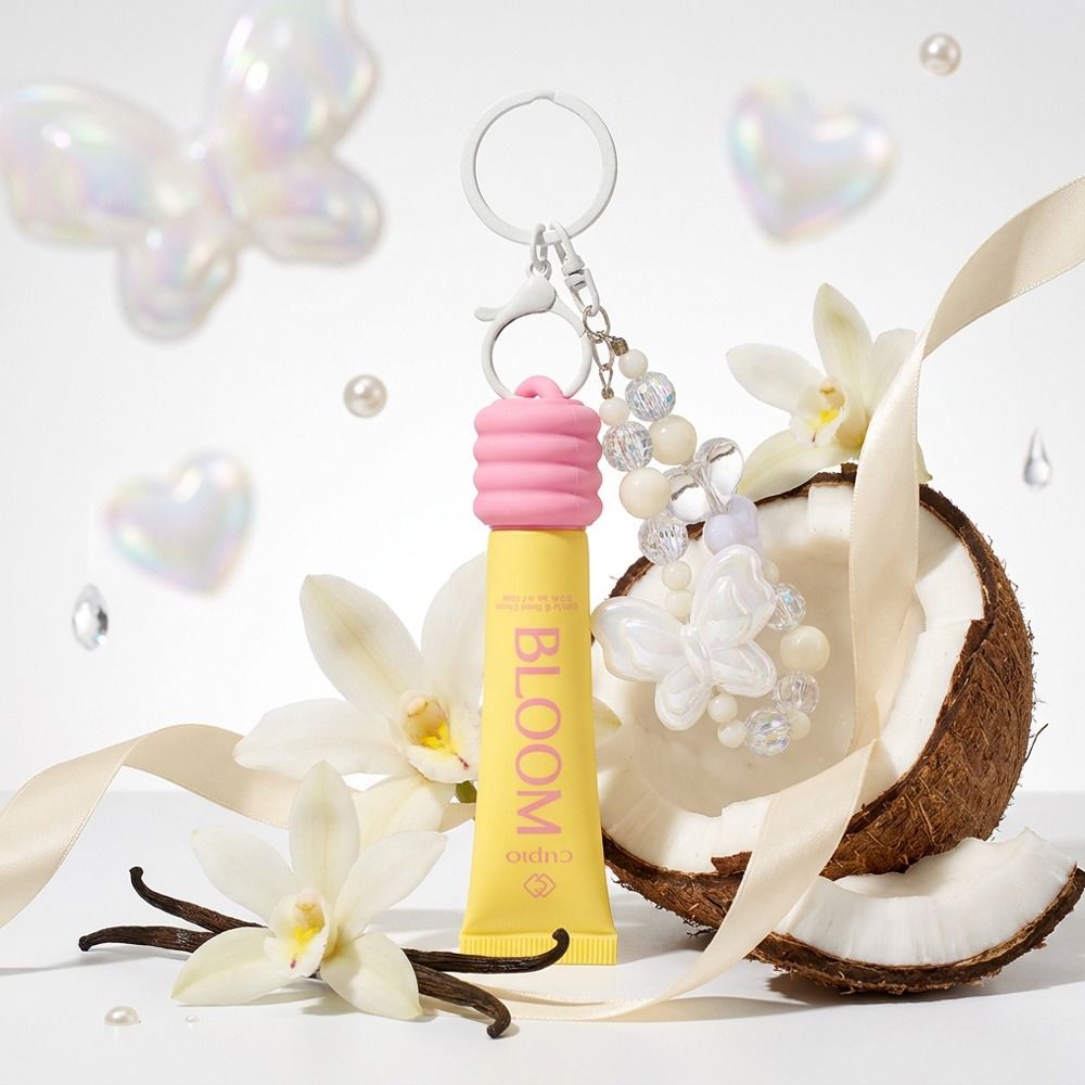 Gelber Cuplo BLOOM Lipgloss als Schlüsselanhänger mit rosa Kappe und Perlenkette, inszeniert mit Kokosnuss, Vanilleschoten und weißen Orchideen