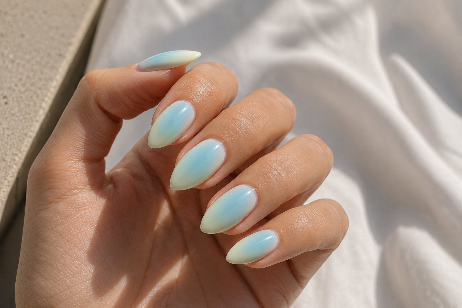 Nahaufnahme mandelförmiger künstlicher Fingernägel mit sanftem Ombré-Verlauf in Hellblau und Weiß auf einer Hand