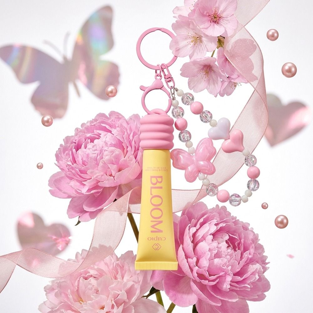 BLOOM Lipgloss in gelber Tube mit rosa Schlüsselanhänger, Perlen und Schleife vor rosa Pfingstrosen, Kirschblüten und Schmetterlingsschatten