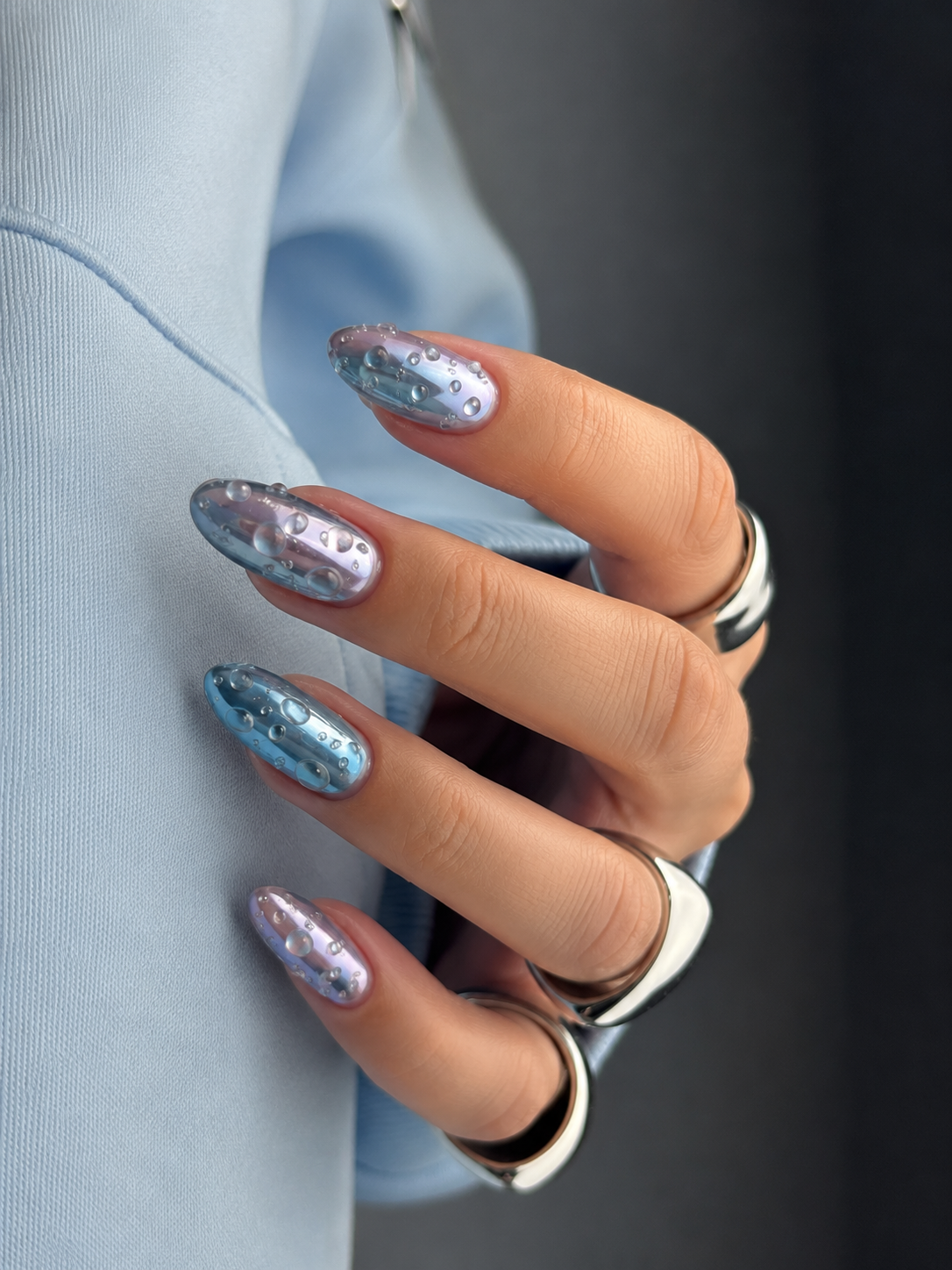 Nahaufnahme einer Hand mit mandelförmigen Kunstnägeln in Blau und Lila mit glänzendem 3D-Wassertropfen-Nailart-Design