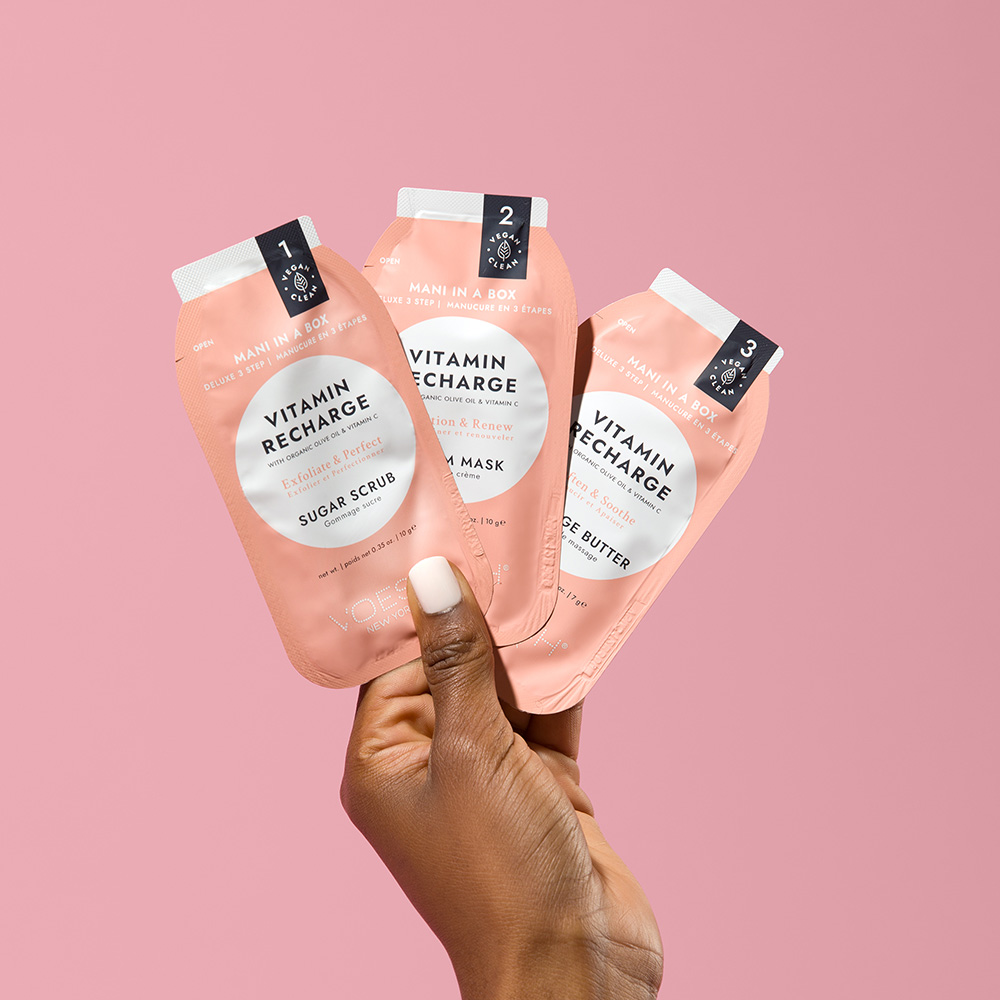 Hand hält das Voesh New York Mani in a Box Vitamin Recharge 3-Step Maniküre-Set mit Sugar Scrub, Mud Mask und Massage Butter vor rosa Hintergrund