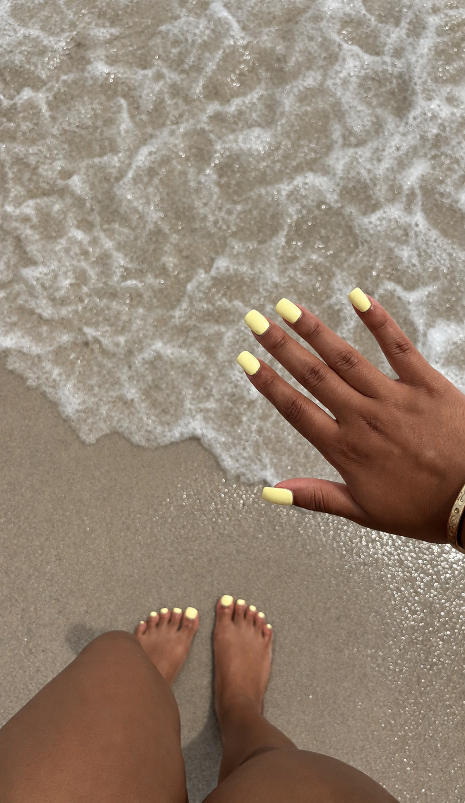 Hand und Füße mit pastellgelbem Nageldesign am Sandstrand, nahe der schäumenden Meereswellen – sommerliche Maniküre und Pediküre