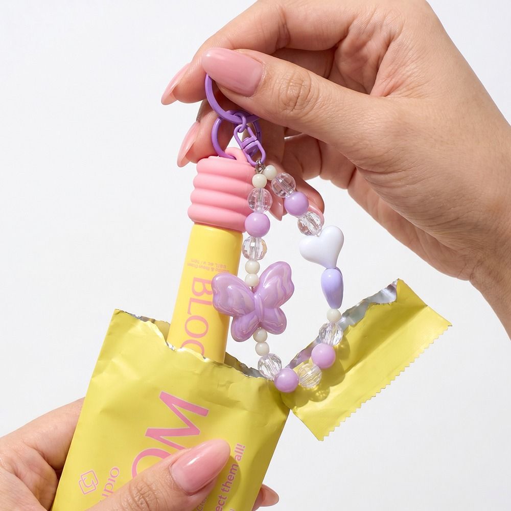 Hand hält gelben Lipgloss mit Schlüsselanhänger aus lila und transparenten Perlen, Schmetterling und Herz als dekoratives Beauty-Accessoire