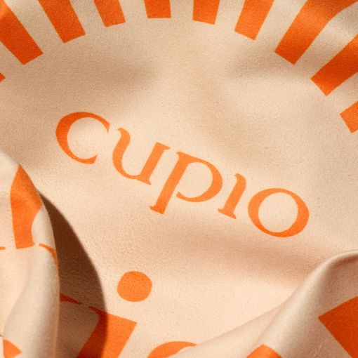 Nahaufnahme eines Stoffes mit dem Aufdruck 'cupio' und grafischen Elementen in leuchtendem Orange auf beigeem Hintergrund
