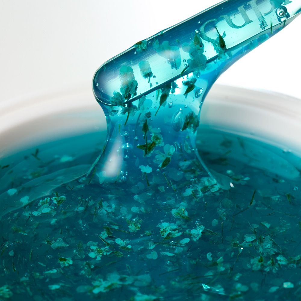 Nahaufnahme eines blauen Gel-Serums mit eingearbeiteten Blütenextrakten auf einem transparenten Spatel – kosmetische Pflege-Textur