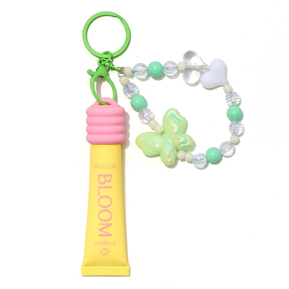 Gelber BLOOM Lipgloss als Schlüsselanhänger mit grünem Ring, rosa Halterung, Perlenarmband und schimmerndem Schmetterlingsanhänger in Pastellfarben