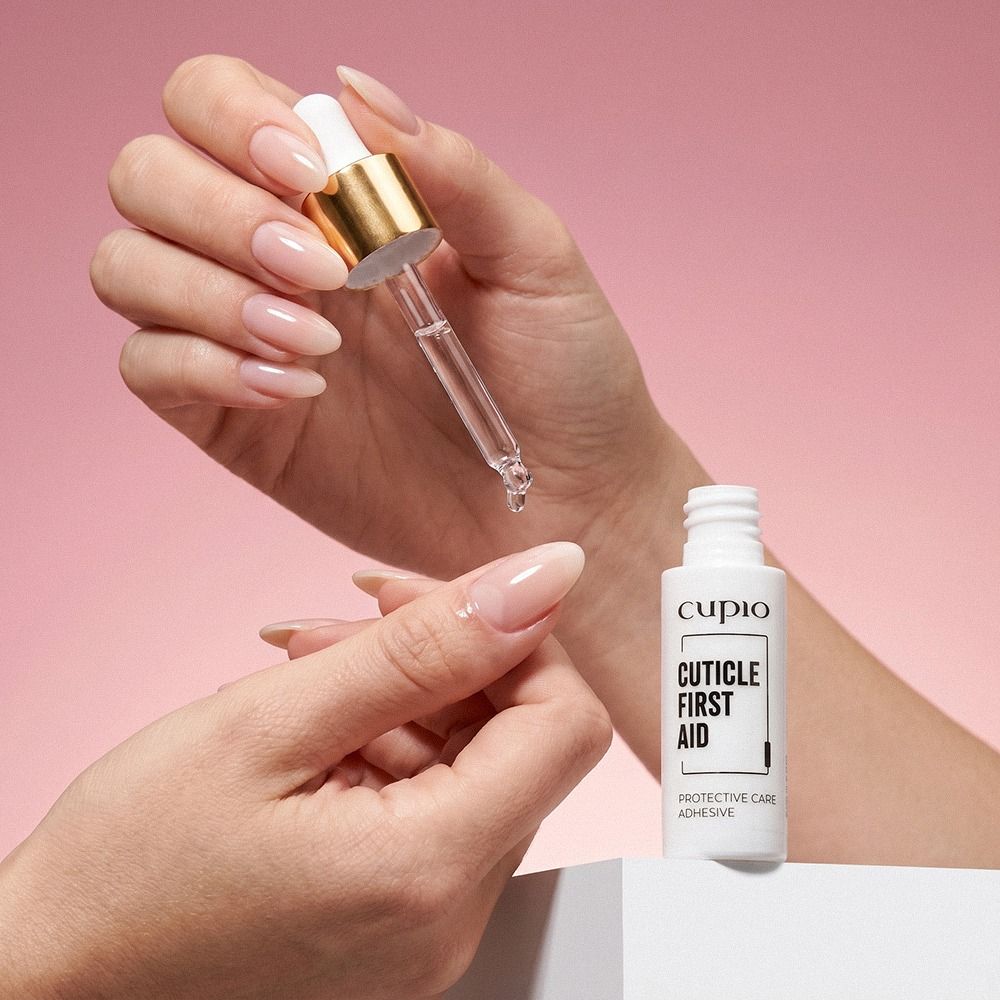 Hand trägt mit Pipette das Cupio Cuticle First Aid Protective Care Adhesive auf die Nagelhaut auf, Nahaufnahme vor rosa Hintergrund