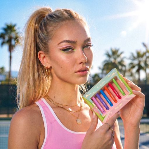 Frau mit Pferdeschwanz hält ein buntes Clinique Chubby Sticks Lippenstift-Set in der Hand, outdoor bei sonnigem Wetter mit Palmen im Hintergrund