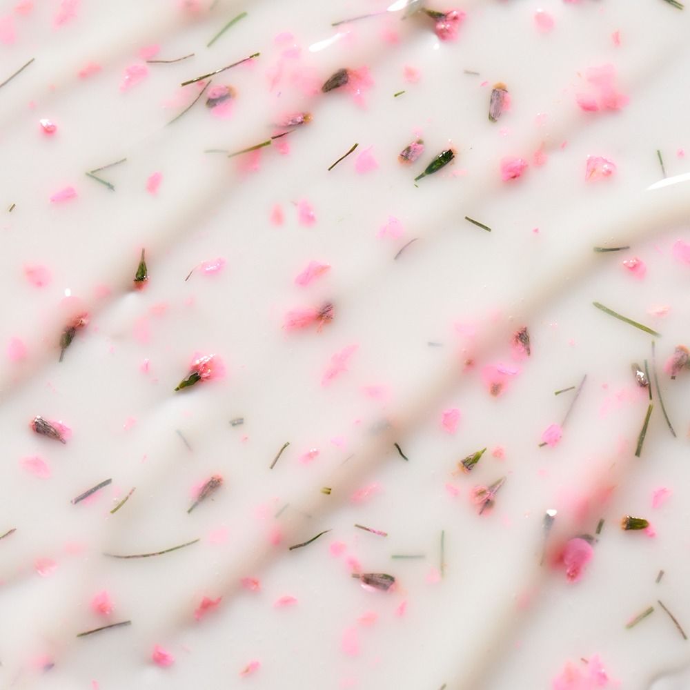 Makroaufnahme einer weißen Duftkerze mit rosa Blütenblättern und feinen Kräutern in dekorativ strukturierter Wachsoberfläche