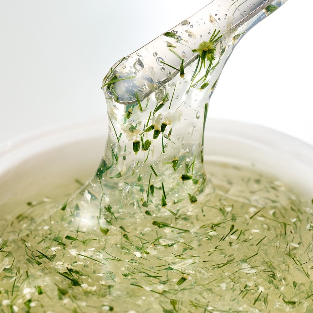 Nahaufnahme eines transparenten Pflegegels mit eingeschlossenen Kräutern und Blüten auf einem Spatel, ideal zur Darstellung natürlicher Kosmetik
